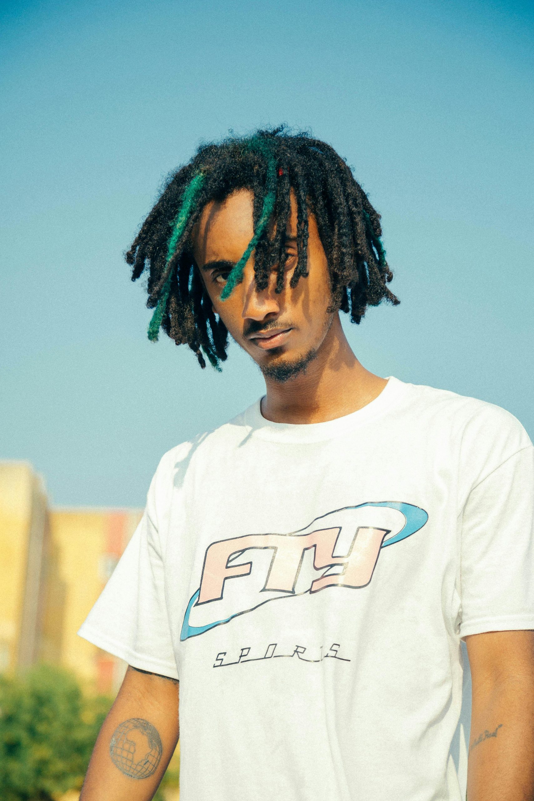 Close-up portrait of a young man with dreadlocks wearing a casual white shirt in daylight.